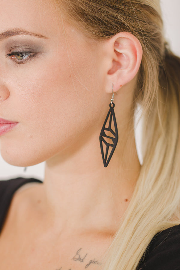 zoom, vue gros plan, paire de boucles d'oreilles graphique, couleur noir. Portées par mannequin Dorine Gaudin. Bijoux fantaisies en silicone, marque française, créateurs, designers. Style pendante, minimaliste, géométrique, losange, triangle. Attache dormeuse en acier inoxydable. Minimisant les allergies, résistant à l'eau/à la chaleur/à la lumière, souple, léger. Cadeau original.