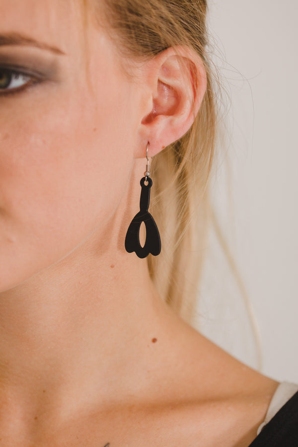 zoom, vue gros plan, paire de boucles d'oreilles clochette, couleur noir. Portées par mannequin Dorine Gaudin. Bijoux fantaisies en silicone, marque française, créateurs, designers. Style nature, fleur, floral, pendante, courte, feuille. Attache dormeuse en acier inoxydable. Minimisant les allergies, résistant à l'eau/à la chaleur/à la lumière, souple, léger. Cadeau original.