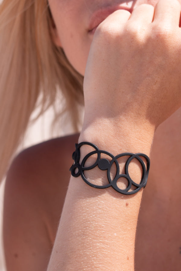 zoom, vue gros plan, bracelet disco, couleur noir. Vue taille réelle, porté sur poignet par mannequin Dorine Gaudin. Bijoux fantaisies en silicone, ladygum, marque française, créateurs, designers. Style manchette femme, rond, cercle, bulle, année 80, géométrique, contemporain. Léger, souple, longueur réglable, bracelet ajustable, fermoir en T, résistant à l'eau, à la chaleur, à la lumière, minimisant les allergies. Cadeau original.