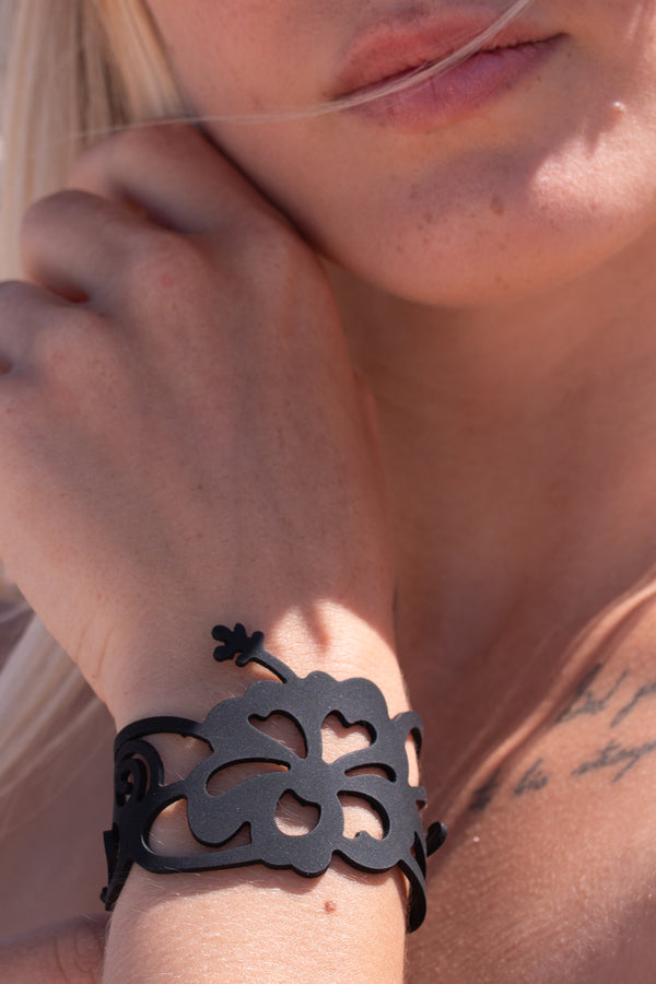 zoom, vue gros plan, bracelet hibiscus, couleur noir. Vue taille réelle, porté sur poignet par mannequin Dorine Gaudin. Bijoux fantaisies en silicone, ladygum, marque française, créateurs, designers. Style manchette femme, nature, fleur, floral, exotique, tropical, île, feuilles, plage. Léger, souple, longueur réglable, bracelet ajustable, fermoir en T, résistant à l'eau, à la chaleur, à la lumière, minimisant les allergies. Cadeau original.
