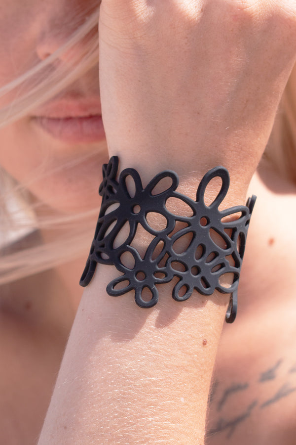 zoom, vue gros plan, bracelet hippie, couleur noir. Vue taille réelle, porté sur poignet par mannequin Dorine Gaudin. Bijoux fantaisies en silicone, ladygum, marque française, créateurs, designers. Style manchette femme, nature, fleur, floral, paquerette, marguerite, année 70. Léger, souple, longueur réglable, bracelet ajustable, fermoir en T, résistant à l'eau, à la chaleur, à la lumière, minimisant les allergies. Cadeau original.