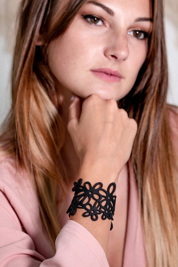 vue gros plan, taille réelle, porté par mannequin sur poignet, youna, bracelet hippie, couleur noir. Silicone, bijoux fantaisies, ladygum, marque française, créateurs, designers. Style manchette, fleur, nature, été, champêtre, plage, marguerite, paquerette, bohème, année 70. Souple, léger, résistant à l'eau, à la chaleur, à la lumière. Longueur réglable, bracelet ajustable. Fermoir en T, plusieurs trou pour régler à la bonne taille.