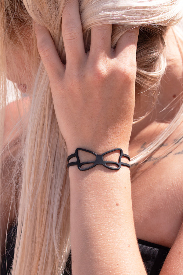 zoom, vue gros plan, bracelet noeud, couleur noir. Vue taille réelle, porté sur poignet par mannequin Dorine Gaudin. Bijoux fantaisies en silicone, ladygum, marque française, créateurs, designers. Style simple, minimaliste, noeud papillon, ruban, manchette femme. Léger, souple, longueur réglable, bracelet ajustable, fermoir en T, résistant à l'eau, à la chaleur, à la lumière, minimisant les allergies. Cadeau original.