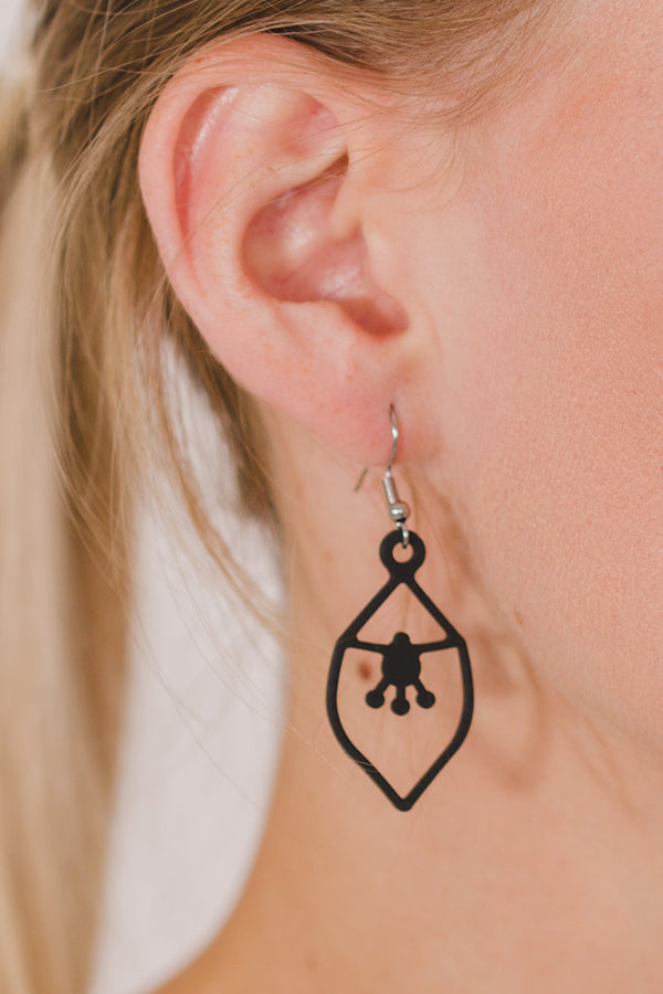 zoom, vue gros plan, boucles d'oreilles Nymphéa, couleur noir, portées sur mannequin Dorine. Attache dormeuse en acier inoxydable. Bijoux fantaisies, marque française, créateurs, designers. Petit modèle, style minimaliste, losange, pampilles, fleurs, géométrique. Silicone, souple, léger, minimisant les allergies, résistant à l'eau/à la chaleur/à la lumière. Idée cadeau original.