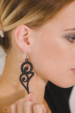 vue gros plan, paire de boucles d'oreilles courbe, grand modèle, couleur noir. Portées par mannequin Dorine Gaudin. Bijoux fantaisies en silicone, marque française, créateurs, designers. Style fleur, floral, rond, longues, pendantes. Attache dormeuse en acier inoxydable. Minimisant les allergies, résistant à l'eau/à la chaleur/à la lumière, souple, léger. Cadeau original.