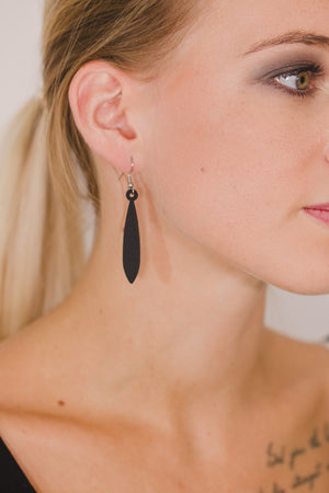 zoom, vue gros plan, paire de boucles d'oreilles emeraude, couleur noir. Portées par mannequin Dorine Gaudin. Bijoux fantaisies en silicone, marque française, créateurs, designers. Style pendante, minimaliste, feuille, longues. Attache dormeuse en acier inoxydable. Minimisant les allergies, résistant à l'eau/à la chaleur/à la lumière, souple, léger. Cadeau original.