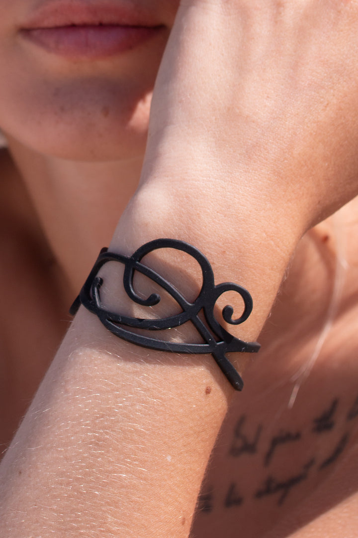 zoom, vue gros plan, bracelet courbe, couleur noir. Vue taille réelle, porté sur poignet par mannequin Dorine Gaudin. Bijoux fantaisies en silicone, ladygum, marque française, créateurs, designers. Style nature, manchette femme, fleur, rond, cercle, tourbillon, abstrait, contemporain. Léger, souple, longueur réglable, bracelet ajustable, fermoir en T, résistant à l'eau, à la chaleur, à la lumière, minimisant les allergies. Cadeau original.