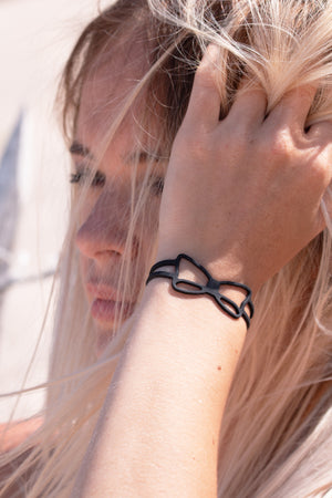 zoom, vue gros plan, bracelet noeud, couleur noir. Vue taille réelle, porté sur poignet par mannequin Dorine Gaudin. Bijoux fantaisies en silicone, ladygum, marque française, créateurs, designers. Style simple, minimaliste, noeud papillon, ruban, manchette femme. Léger, souple, longueur réglable, bracelet ajustable, fermoir en T, résistant à l'eau, à la chaleur, à la lumière, minimisant les allergies. Cadeau original.