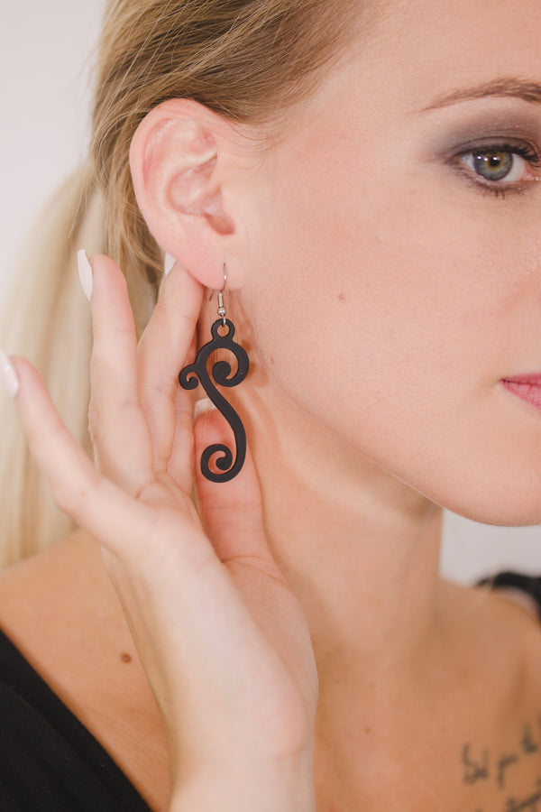 zoom, vue gros plan, paire de boucles d'oreilles courbe, petit modèle, couleur noir. Portées par mannequin Dorine Gaudin. Bijoux fantaisies en silicone, marque française, créateurs, designers. Style fleur, floral, rond, longues, pendantes. Attache dormeuse en acier inoxydable. Minimisant les allergies, résistant à l'eau/à la chaleur/à la lumière, souple, léger. Cadeau original.